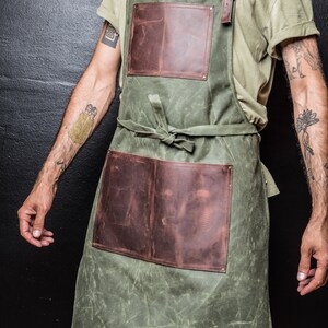 May include: A green canvas apron with two brown leather pockets and adjustable straps. The apron is tied at the waist with a brown leather strap.