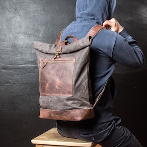 May include: A charcoal gray roll-top backpack featuring a brown leather base and a matching brown leather front pocket with a zippered closure. The bag includes a top handle, adjustable shoulder straps, and antique brass hardware accents.