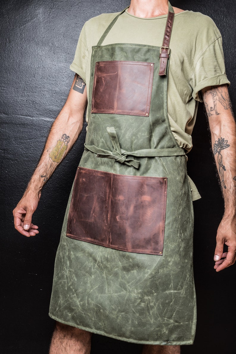 May include: A green canvas apron with two brown leather pockets and adjustable straps. The apron is tied at the waist with a brown leather strap.
