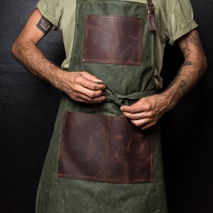 May include: A green canvas apron with brown leather accents and pockets. The apron has adjustable straps and a tie at the waist.