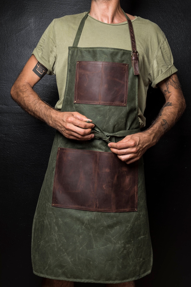 May include: A green canvas apron with brown leather accents and pockets. The apron has adjustable straps and a tie at the waist.