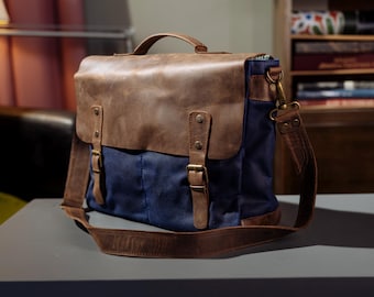 May include: A navy blue canvas messenger bag featuring a brown leather flap, a top handle, and an adjustable shoulder strap. The bag includes two vertical leather straps with metal buckles and rivets, securing the front flap over the main compartment.