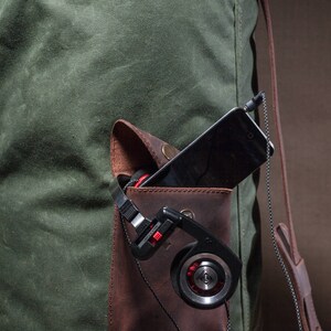 May include: A green canvas bag with a brown leather pocket. The pocket contains a black smartphone and a pair of black headphones with red accents.
