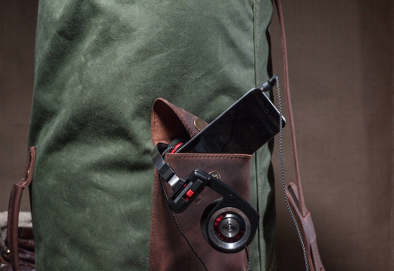 May include: A green canvas bag with a brown leather pocket. The pocket contains a black smartphone and a pair of black headphones with red accents.