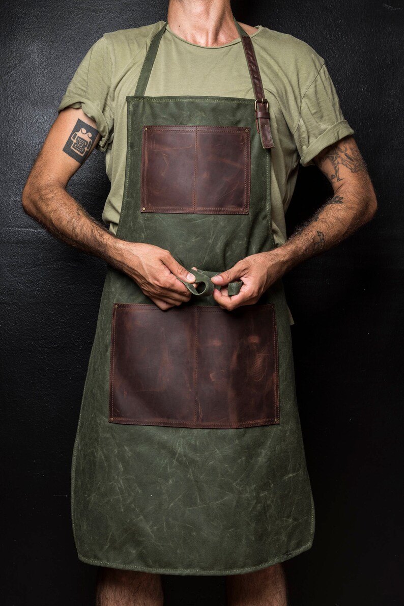 May include: A green canvas apron with two large brown leather pockets. The apron has adjustable straps and a leather buckle.
