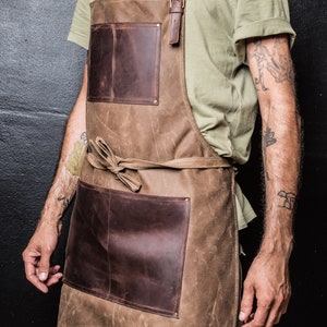 May include: A brown leather and canvas apron with two large pockets and adjustable straps. The apron is tied at the waist with a brown leather strap.