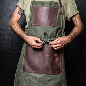 May include: A green canvas apron with brown leather straps and pockets. The apron has a simple design and is tied at the waist.