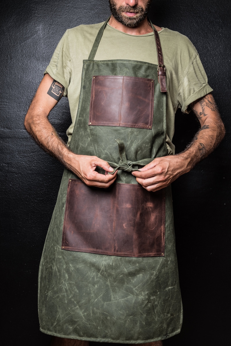 May include: A green canvas apron with brown leather straps and pockets. The apron has a simple design and is tied at the waist.