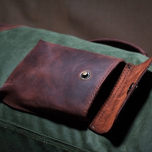 May include: A brown leather pocket with a flap closure and a metal button. The pocket is attached to a green canvas bag.