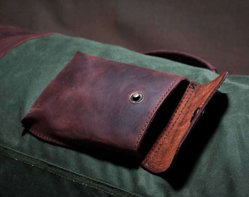May include: A brown leather pocket with a flap closure and a metal button. The pocket is attached to a green canvas bag.