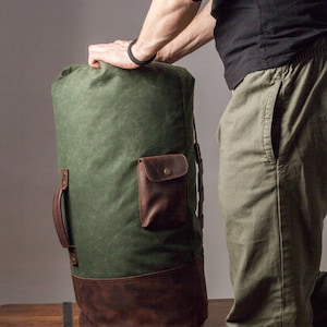 May include: A green canvas duffel bag with brown leather accents. The bag has a large main compartment, a smaller front pocket, and two adjustable straps. The bag is sitting on a wooden surface.