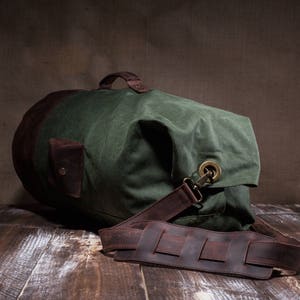 May include: A green canvas duffel bag with brown leather accents and a long adjustable strap. The bag has a large main compartment and a smaller pocket on the front.