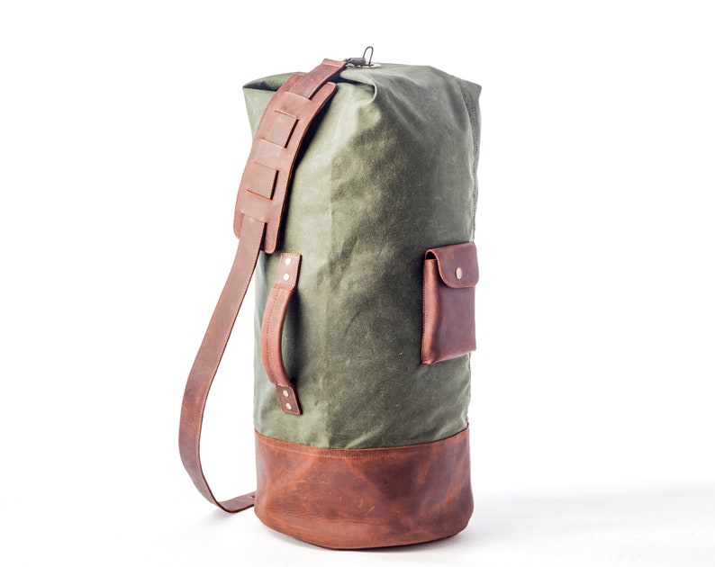 May include: A green canvas duffel bag with brown leather accents and a small pocket on the front. The bag has a long adjustable strap and a top handle.