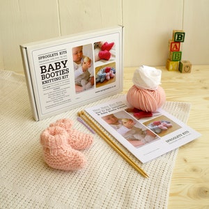 May include: A baby booties knitting kit with a pair of pink knitted booties, yarn, knitting needles, and instructions. The kit box reads "Baby Booties Knitting Kit" and includes images of finished booties. Wooden blocks spell out "BABY".