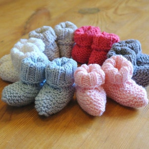 May include: A collection of hand-knitted baby booties in various colors, including white, blue, pink, red, and gray. The booties are arranged on a wooden surface, showcasing their soft texture and cozy design. These knitted baby shoes are perfect for newborns.