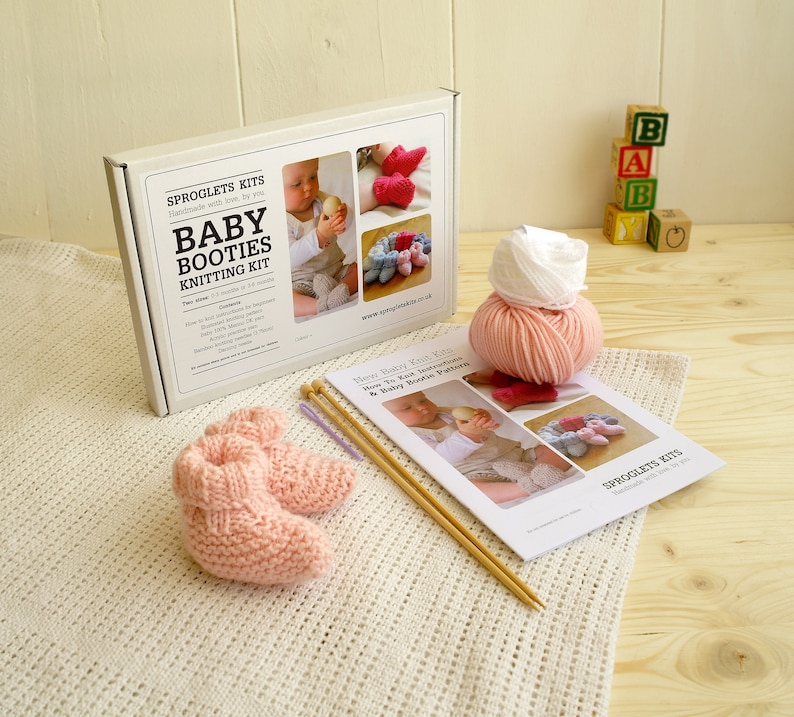 May include: A baby booties knitting kit with a pair of pink knitted booties, yarn, knitting needles, and instructions. The kit box reads "Baby Booties Knitting Kit" and includes images of finished booties. Wooden blocks spell out "BABY".