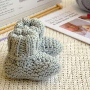 May include: A pair of light blue knitted baby booties. The booties are made of soft yarn and have a ribbed cuff. Knitting needles and a pattern are in the background. The booties are approximately 3 inches long.