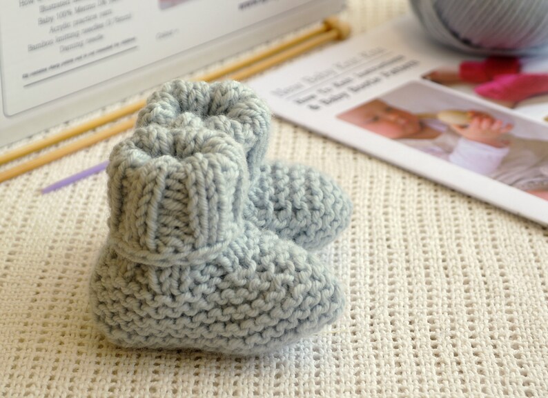 May include: A pair of light blue knitted baby booties. The booties are made of soft yarn and have a ribbed cuff. Knitting needles and a pattern are in the background. The booties are approximately 3 inches long.