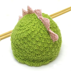Puede incluir: Sombrero de punto verde con una púa de dinosaurio de crochet rosa en la parte superior. El sombrero está hecho con un punto texturizado y está sobre una superficie blanca.