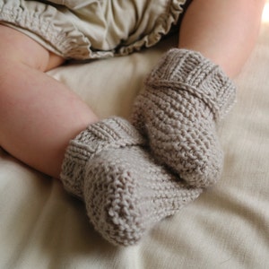May include: A pair of knitted baby booties in a neutral beige color. The booties are made of a textured yarn and have a ribbed cuff. The image shows the booties on a baby's foot, resting on a soft, light-colored fabric.