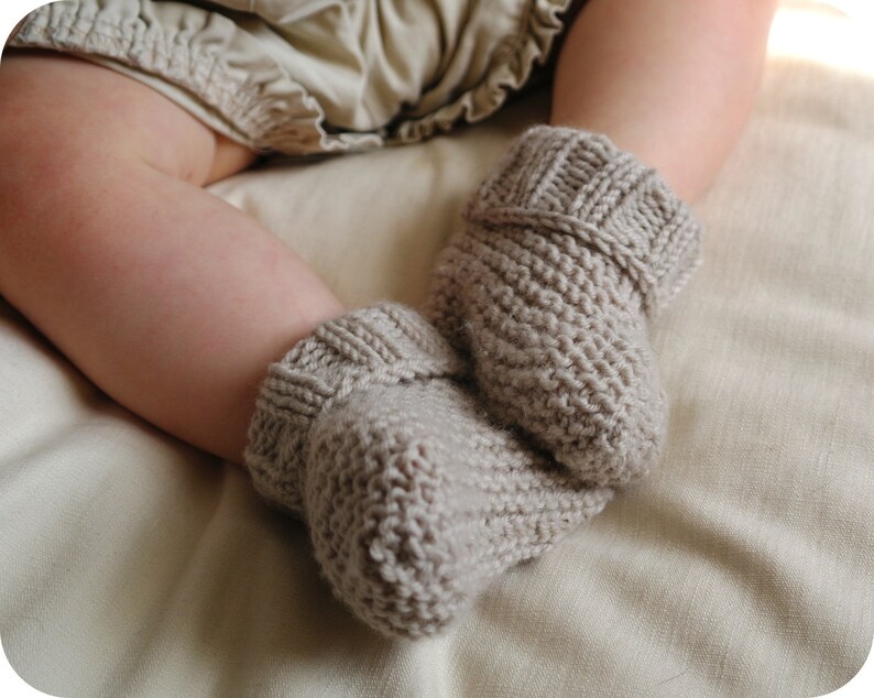 May include: A pair of knitted baby booties in a neutral beige color. The booties are made of a textured yarn and have a ribbed cuff. The image shows the booties on a baby's foot, resting on a soft, light-colored fabric.