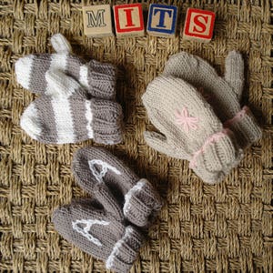 May include: Two pairs of knitted baby mittens. One pair is gray with white letters, the other is beige with pink flower embroidery. The word "MITS" is spelled out with colorful wooden blocks.