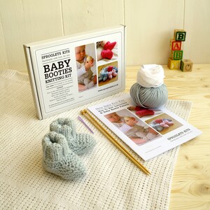 May include: A baby booties knitting kit with a pair of light blue knitted booties, knitting needles, and yarn. The kit includes a box with the text "Baby Booties Knitting Kit" and a pattern. Wooden blocks spell out "BAB".