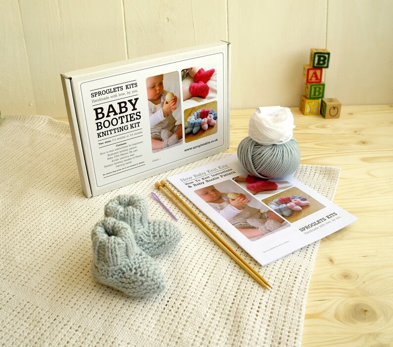 May include: A baby booties knitting kit with a pair of light blue knitted booties, knitting needles, and yarn. The kit includes a box with the text "Baby Booties Knitting Kit" and a pattern. Wooden blocks spell out "BAB".
