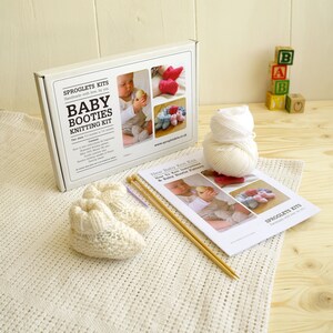 May include: A baby booties knitting kit with a white box, yarn, knitting needles, and a pattern. The box reads "Baby Booties Knitting Kit" and includes images of finished booties. A pair of cream-colored knitted booties sits on a white blanket.