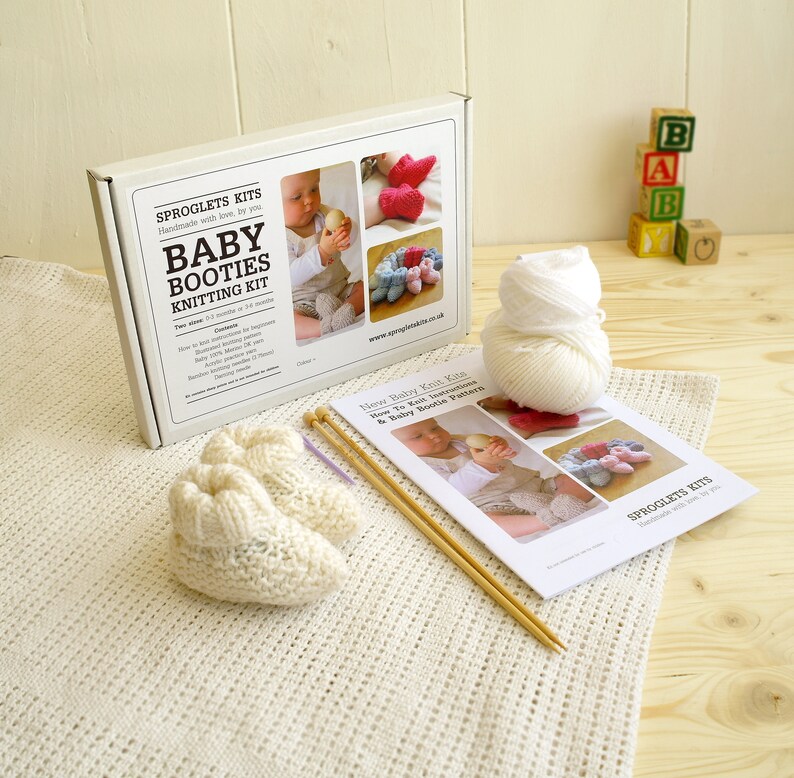 May include: A baby booties knitting kit with a white box, yarn, knitting needles, and a pattern. The box reads "Baby Booties Knitting Kit" and includes images of finished booties. A pair of cream-colored knitted booties sits on a white blanket.