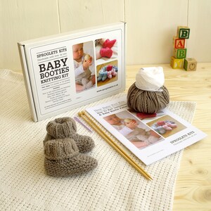 May include: A baby booties knitting kit with a pair of knitted booties, yarn, knitting needles, and instructions. The kit includes a box with the text "Baby Booties Knitting Kit" and images of finished booties. The booties are a light brown color.