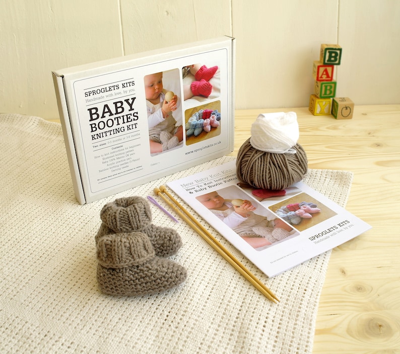 May include: A baby booties knitting kit with a pair of knitted booties, yarn, knitting needles, and instructions. The kit includes a box with the text "Baby Booties Knitting Kit" and images of finished booties. The booties are a light brown color.