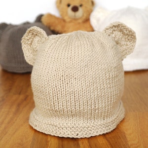 Pode incluir: Um chapéu de bebé de malha bege com orelhas de urso. O chapéu é feito com um fio macio e texturizado e tem um design simples e clássico.