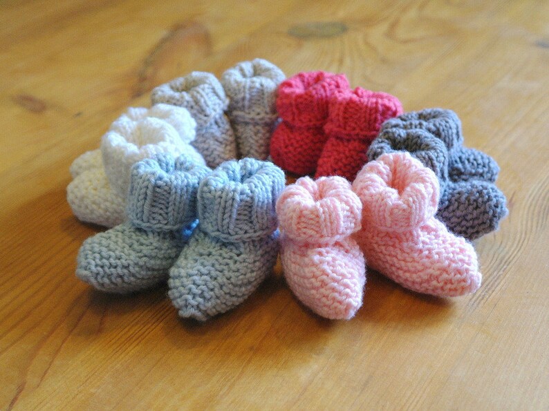 May include: A collection of hand-knitted baby booties in various colors, including white, blue, pink, red, and gray. The booties are arranged on a wooden surface, showcasing their soft texture and cozy design. These knitted baby shoes are perfect for newborns.