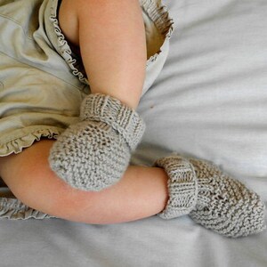 May include: A pair of gray knitted baby booties. The booties are made of a soft, textured yarn and have a ribbed cuff. The baby's legs are visible, resting on a light gray surface.