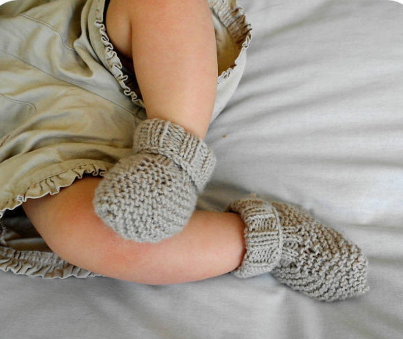 May include: A pair of gray knitted baby booties. The booties are made of a soft, textured yarn and have a ribbed cuff. The baby's legs are visible, resting on a light gray surface.