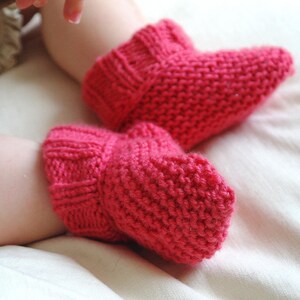 May include: A pair of bright pink knitted baby booties. The booties are made of a thick yarn and have a ribbed cuff. The booties are designed to keep a baby's feet warm and cozy. The booties are a vibrant shade of pink.