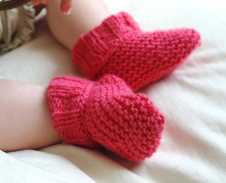 May include: A pair of bright pink knitted baby booties. The booties are made of a thick yarn and have a ribbed cuff. The booties are designed to keep a baby's feet warm and cozy. The booties are a vibrant shade of pink.