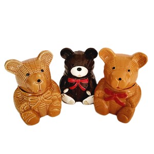 May include: Three ceramic teddy bear jars in shades of brown and dark brown. Each bear has a bow and a lid. The jars are on a white background. The bears are a decorative item.