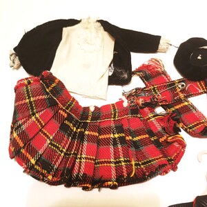 May include: A collection of doll clothing, including a red and black plaid pleated skirt, a black jacket, a white shirt with lace trim, a black beret, and a plaid scarf. The items are arranged on a white surface.