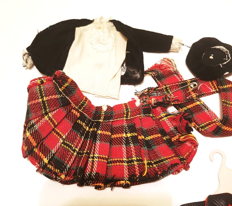 May include: A collection of doll clothing, including a red and black plaid pleated skirt, a black jacket, a white shirt with lace trim, a black beret, and a plaid scarf. The items are arranged on a white surface.