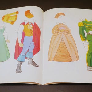 Pode incluir: Livro aberto com bonecas de papel de v&aacute;rias roupas. Inclui um vestido verde, um casaco amarelo, um pr&iacute;ncipe com capa vermelha, uma princesa com vestido amarelo e um uniforme militar verde. As roupas s&atilde;o para recortar e brincar.
