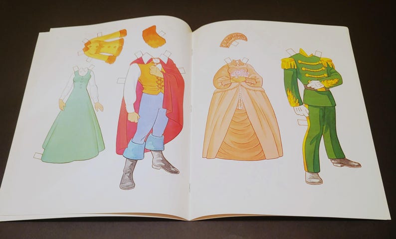 Pode incluir: Livro aberto com bonecas de papel de v&aacute;rias roupas. Inclui um vestido verde, um casaco amarelo, um pr&iacute;ncipe com capa vermelha, uma princesa com vestido amarelo e um uniforme militar verde. As roupas s&atilde;o para recortar e brincar.