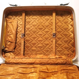 May include: Open vintage suitcase with a gold quilted interior. The interior features a zippered pocket, two straps, and a fold-out section. The exterior is silver with a white handle and clasps.