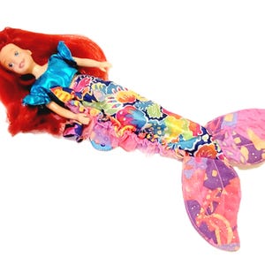 May include: A red-haired mermaid doll wearing a blue and turquoise top and a colourful, floral tail. The tail has a pink and purple fin.