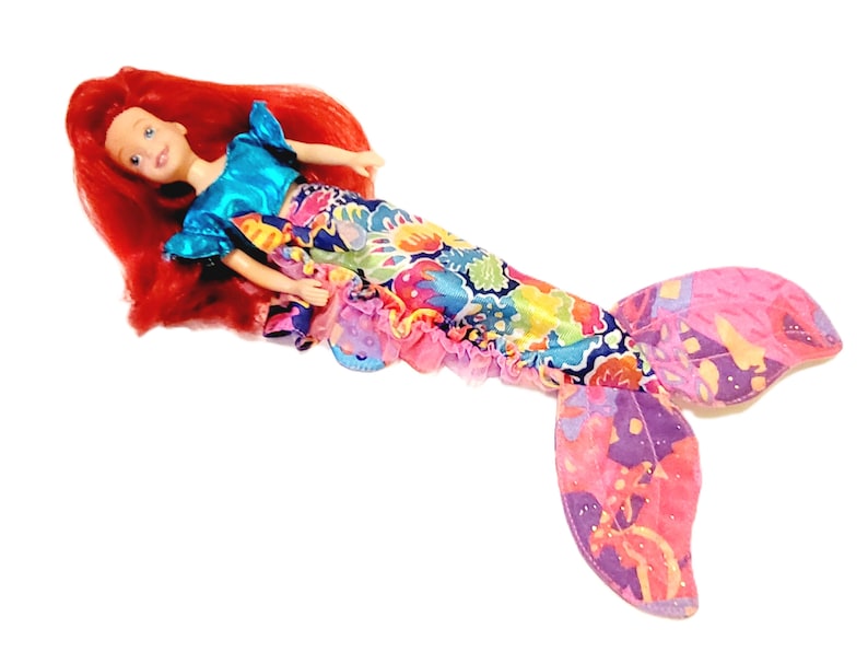 May include: A red-haired mermaid doll wearing a blue and turquoise top and a colourful, floral tail. The tail has a pink and purple fin.