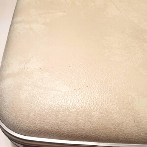 May include: Close-up of a vintage suitcase with a cream-colored, textured exterior. The suitcase has a silver metal trim and shows signs of wear, including discoloration and minor blemishes. The overall aesthetic suggests a classic travel item.