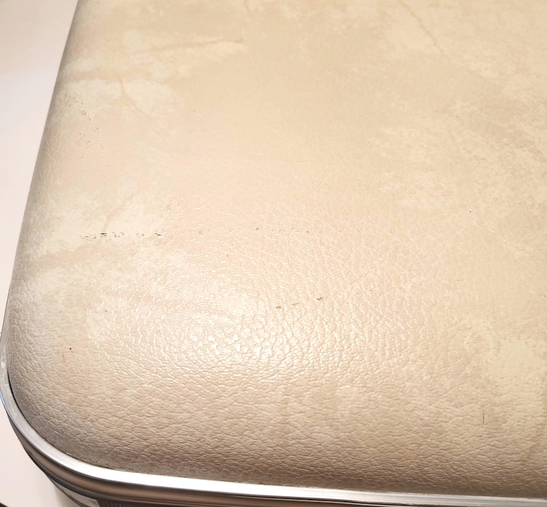May include: Close-up of a vintage suitcase with a cream-colored, textured exterior. The suitcase has a silver metal trim and shows signs of wear, including discoloration and minor blemishes. The overall aesthetic suggests a classic travel item.