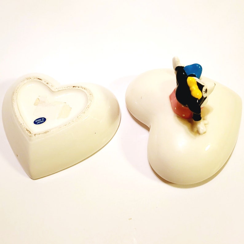 May include: White ceramic heart-shaped trinket box with a cartoon character figurine on top. The figurine is black and white with yellow and blue accents. The box is made in Japan.