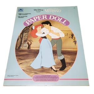 Pode incluir: Um livro de bonecas de papel vintage de "A Pequena Sereia". A capa apresenta Ariel e o Pr&iacute;ncipe Eric em um abra&ccedil;o rom&acirc;ntico. O livro &eacute; azul claro com o t&iacute;tulo "Paper Doll" em um oval rosa. O texto na capa diz "Precut Fashions and 2 Dolls" e "No Scissors Needed."